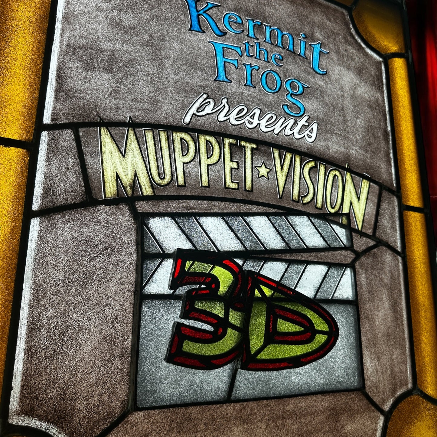 Decorative stained glass panel with 'Kermit the Frog presents Muppet Vision 3D' text against a red curtain with spotlight background
