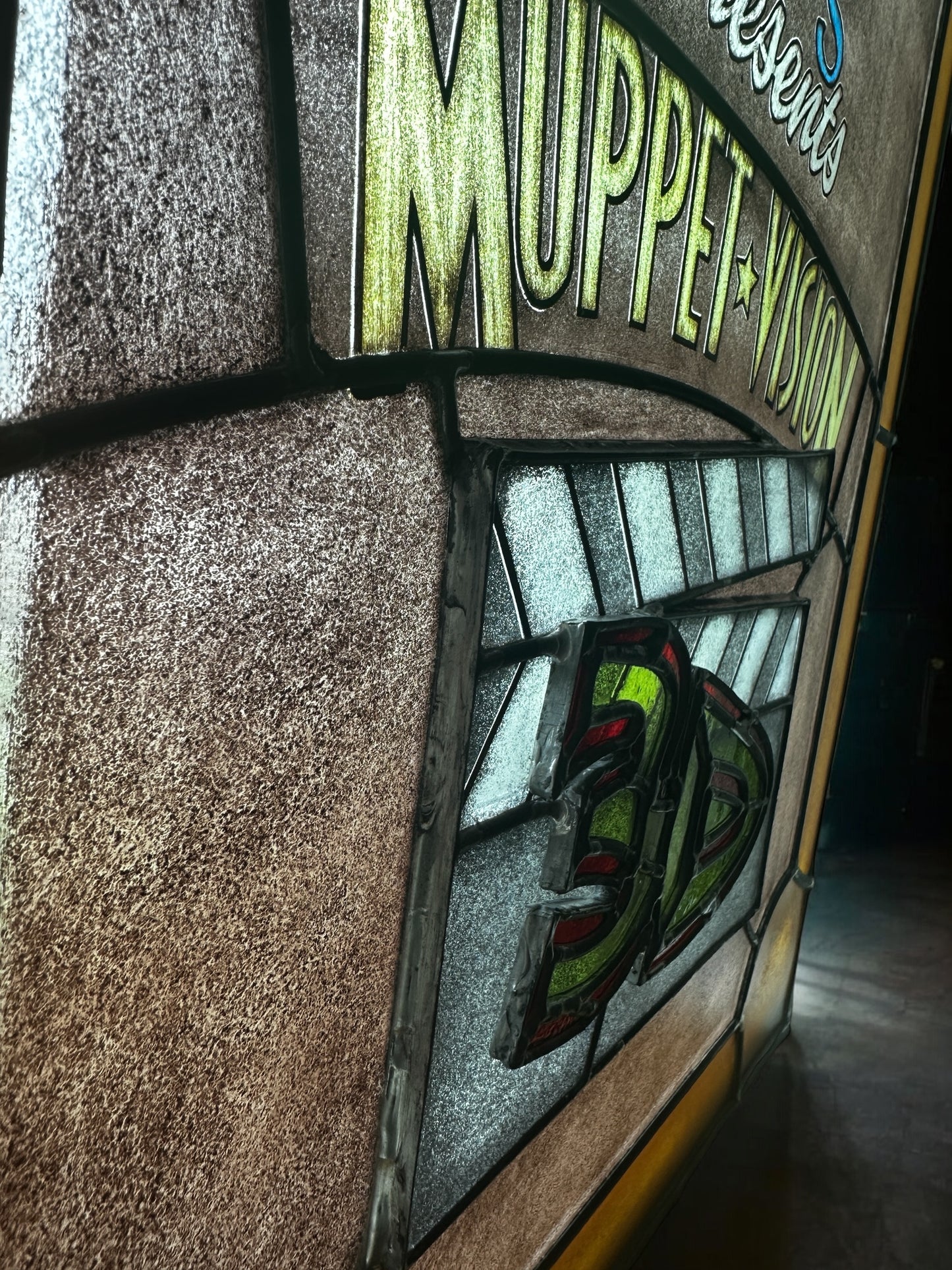 Decorative stained glass panel with 'presents Muppet Vision 3D' text against dark backgorund. '3D' letters are layered off the stained glass panel to to be dimensional