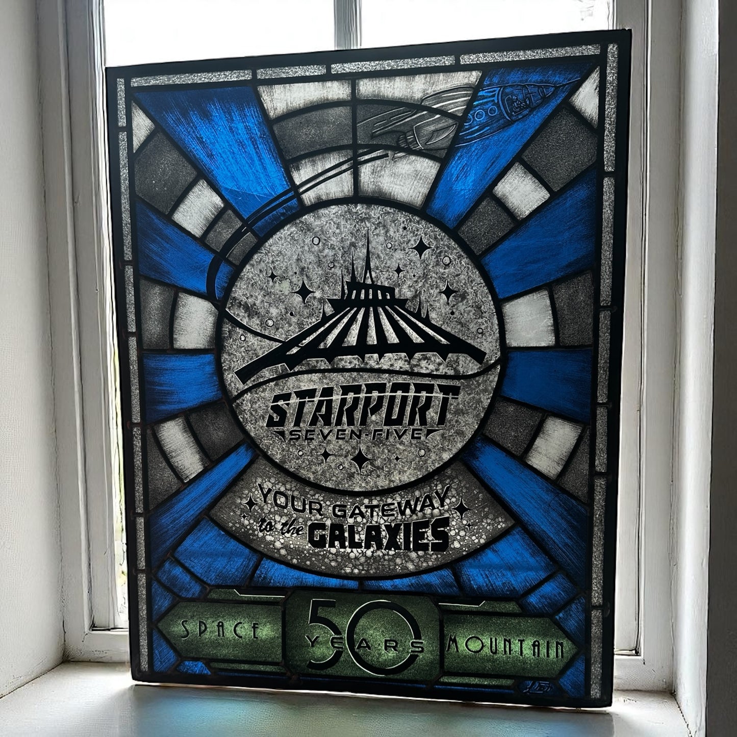 Stylized sign with 'Starport Seven Five' design, stained glass, on a window sill. Blue and white launch tunnel background. '50 years Space Mountain' text. rocket with astronaut blasting off. 'Your Gateway to the Galaxies' text.