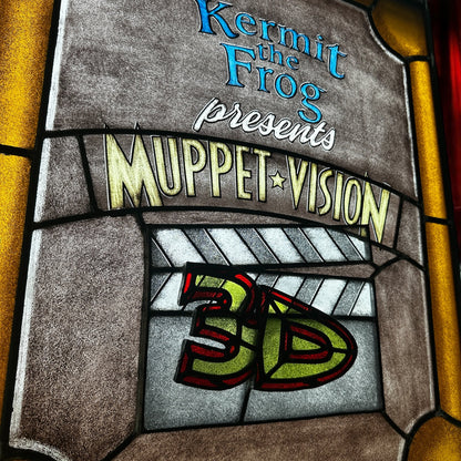 Decorative stained glass panel with 'Kermit the Frog presents Muppet Vision 3D' text against a red curtain with spotlight background